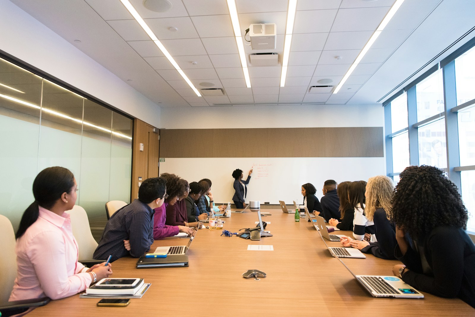 Colleagues seated around a long conference table listen to a woman presenting at the front of the room.