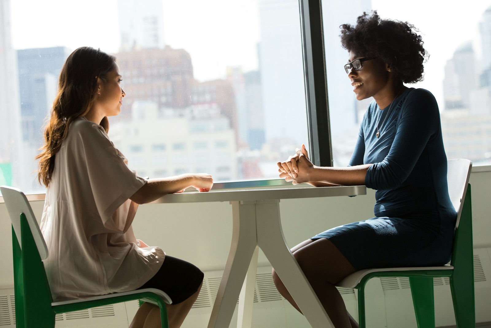 Two women talk across a small table beside an office window.