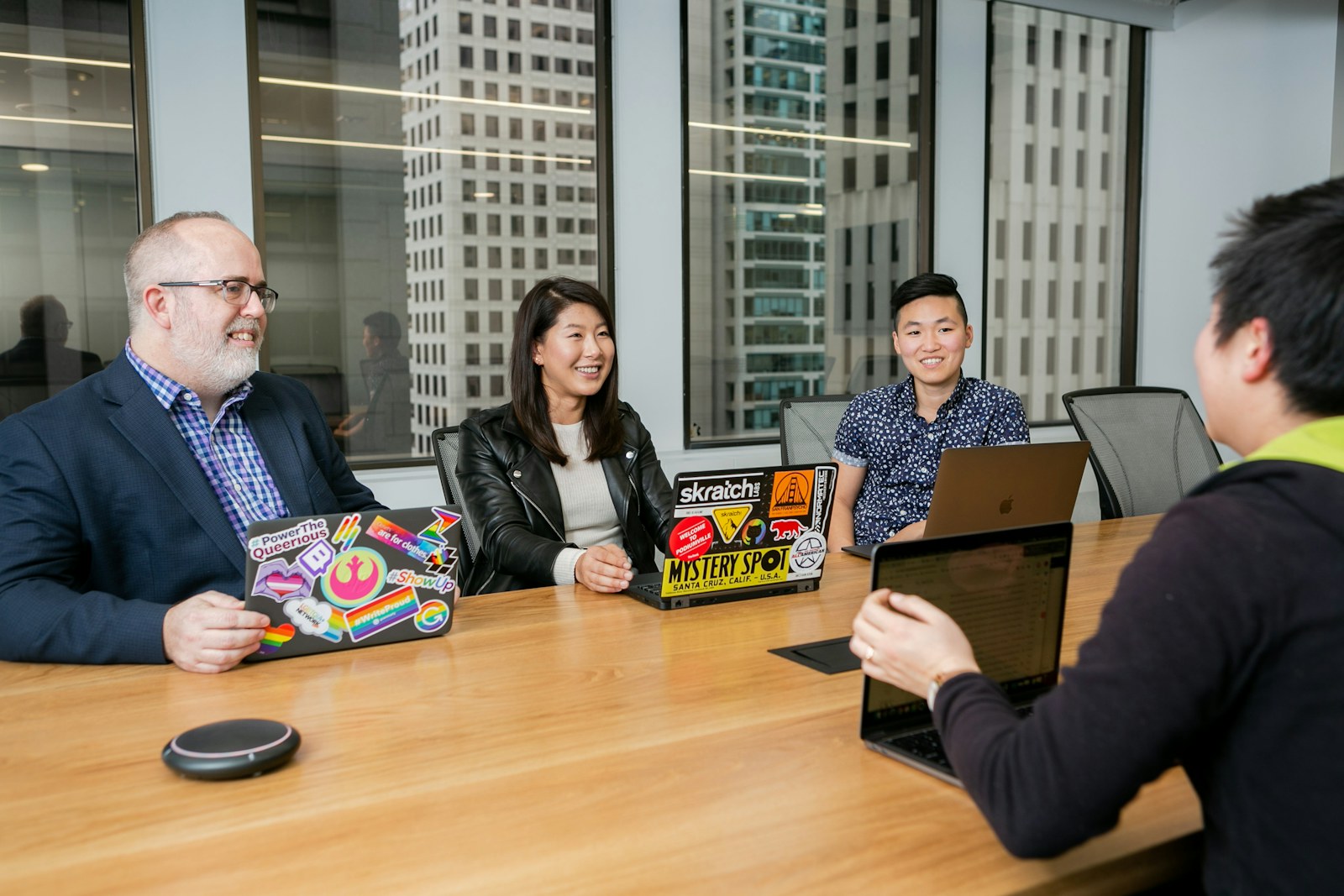 Four colleagues discuss their work around a table with laptops and a large office window.