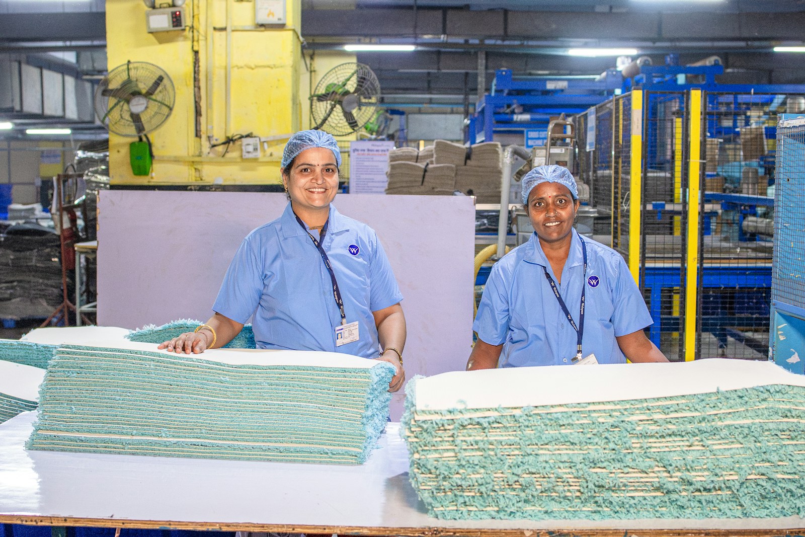 Two workers in blue uniforms and hair coverings stand beside folded textiles on a factory production line.