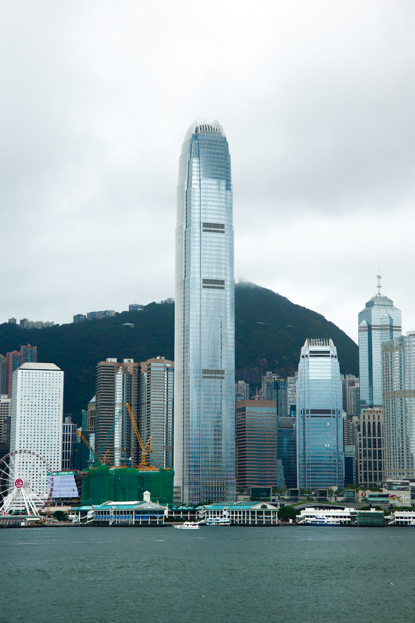 Hong Kong waterfront towers, a Ferris wheel and wooded hills beneath a cloudy sky.